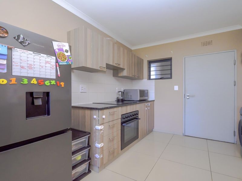 A functional kitchen featuring light wood-finish cabinetry, dark laminate countertops, built-in oven and hob, tiled backsplash, and neutral floor tiles.