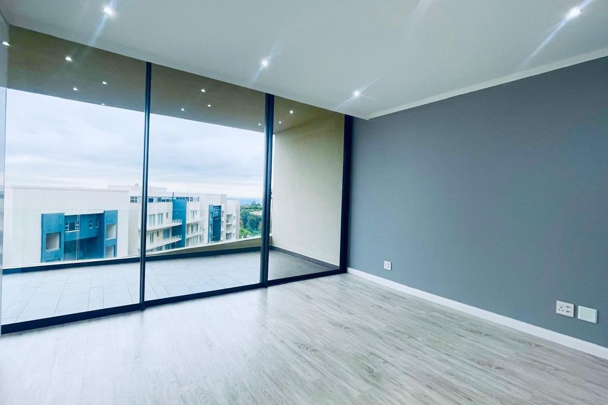A bright, modern living area featuring light wood-look flooring, recessed ceiling lighting, and large floor-to-ceiling glass sliding doors opening onto a private balcony.