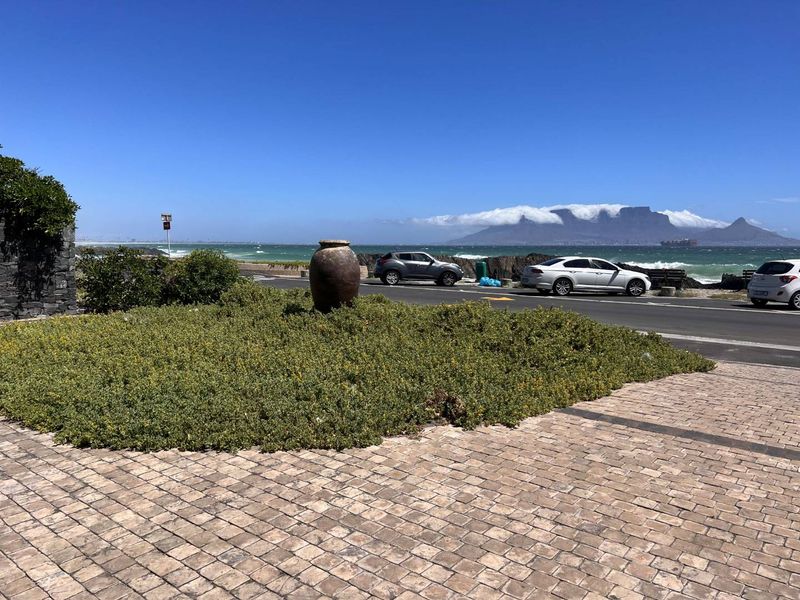 A scenic coastal view overlooking the ocean and Table Mountain from a paved walkway with landscaped garden features.