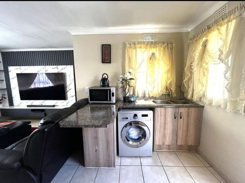 Open-plan living area featuring a compact kitchenette with built-in cabinetry, sink, and laundry appliance space, adjacent to a modern media wall.