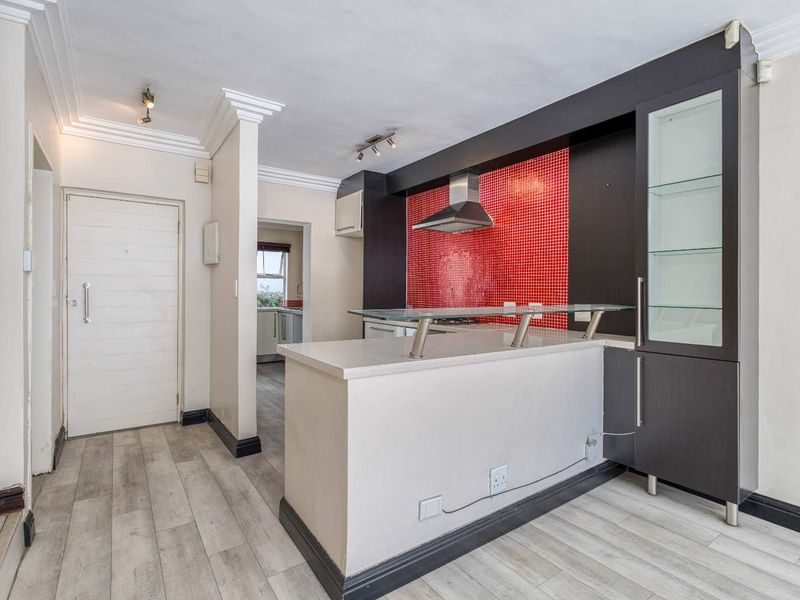 A modern kitchen featuring a breakfast bar, striking red mosaic backsplash, built-in dark cabinetry with glass shelving, and light-toned laminate flooring.