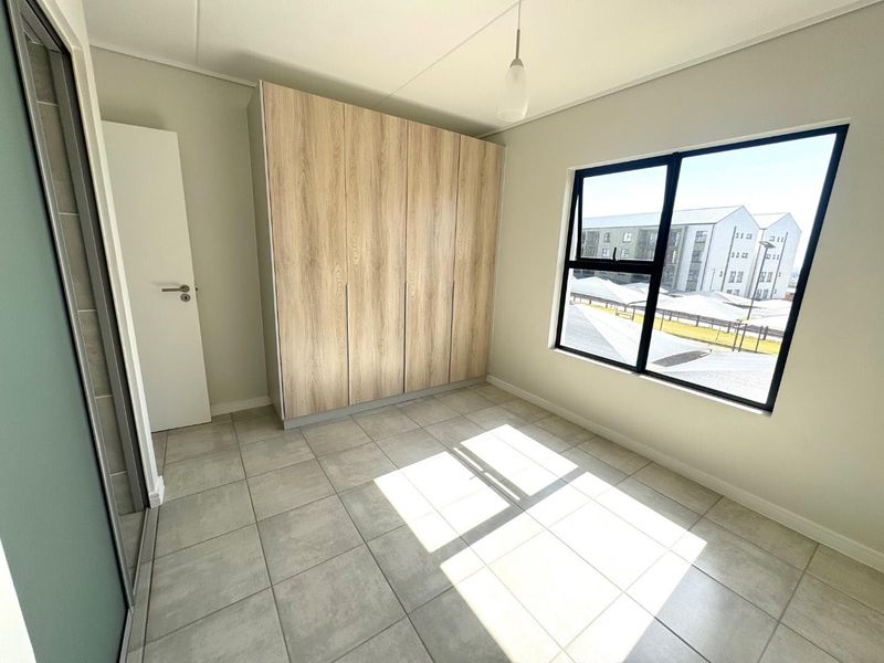 A bright, modern bedroom featuring neutral tiled flooring, built-in wood-finish cupboards, and large aluminium-framed windows providing ample natural light.