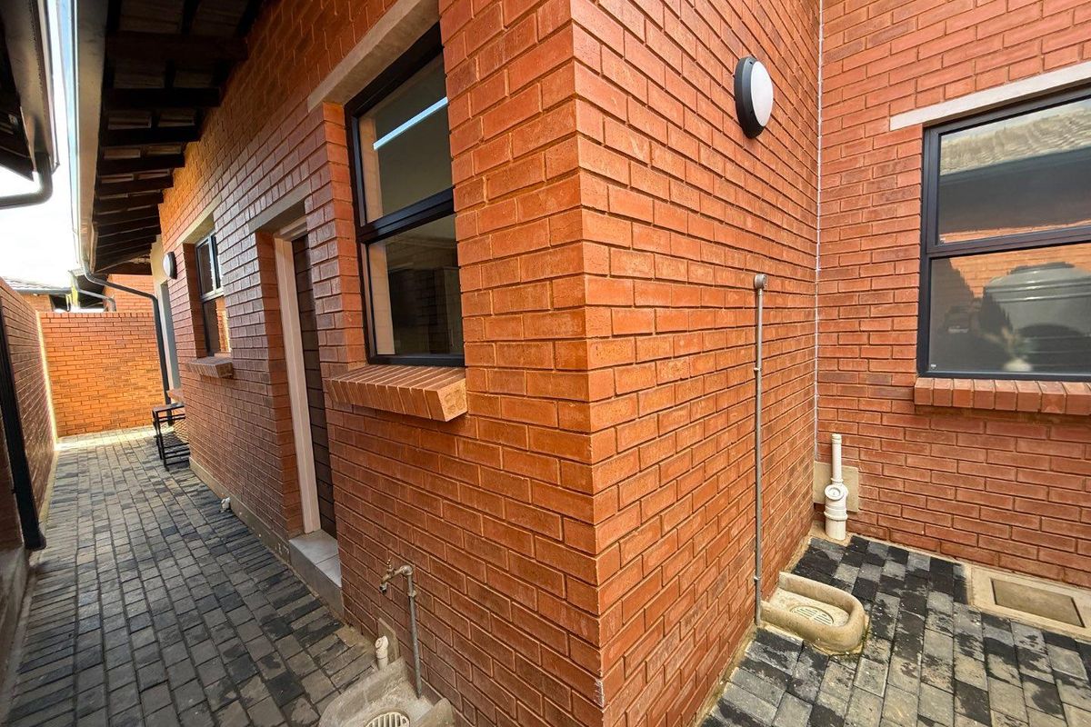 A side exterior view of the townhouse featuring exposed red brickwork, paved walkways, external wall-mounted lighting, and functional drainage points.