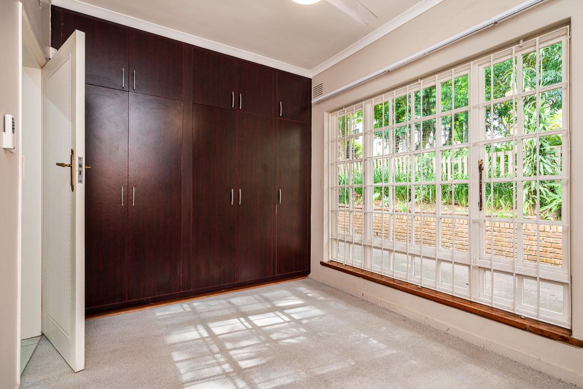 A bright, empty bedroom featuring extensive dark wood built-in cupboards, carpeted flooring, and large windows fitted with security bars.