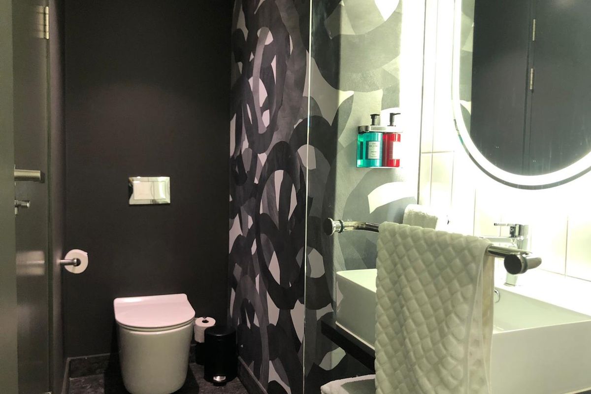 A modern bathroom featuring a wall-mounted toilet, vanity basin with a backlit mirror, and bold patterned wall finishes.