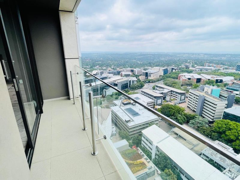 A modern apartment balcony featuring a glass balustrade with stainless steel railings, offering expansive elevated views of the surrounding urban landscape.