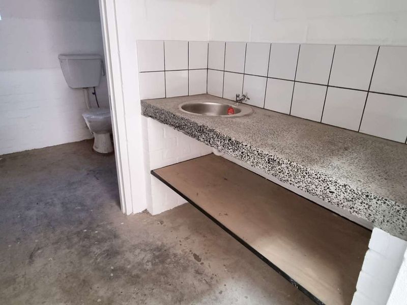 A functional utility area featuring a concrete countertop with an inset sink, tiled splashback, and an adjacent toilet facility with exposed brick walls.