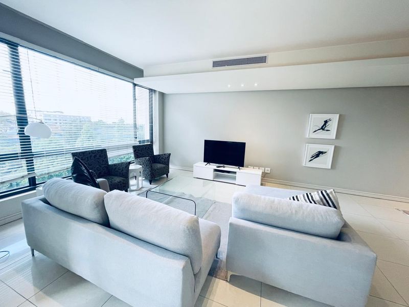 A bright and modern living room featuring large floor-to-ceiling windows, ducted air conditioning, and neutral tiled flooring.
