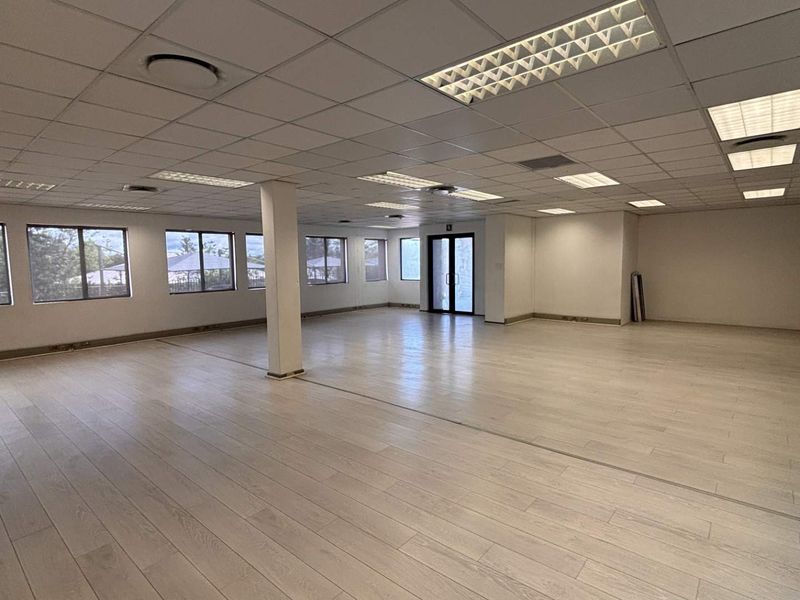 A spacious, open-plan office area featuring light-toned laminate flooring, a suspended ceiling with recessed lighting, and large windows providing natural light.