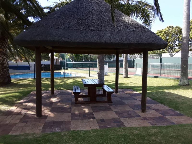 Outdoor communal area featuring a thatched gazebo with paved seating space, overlooking a swimming pool and tennis court facilities.