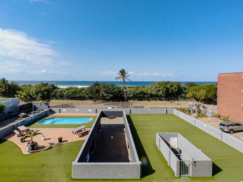 Aerial view of a communal apartment complex pool area featuring artificial turf, paved patio, and perimeter security walls with ocean views.