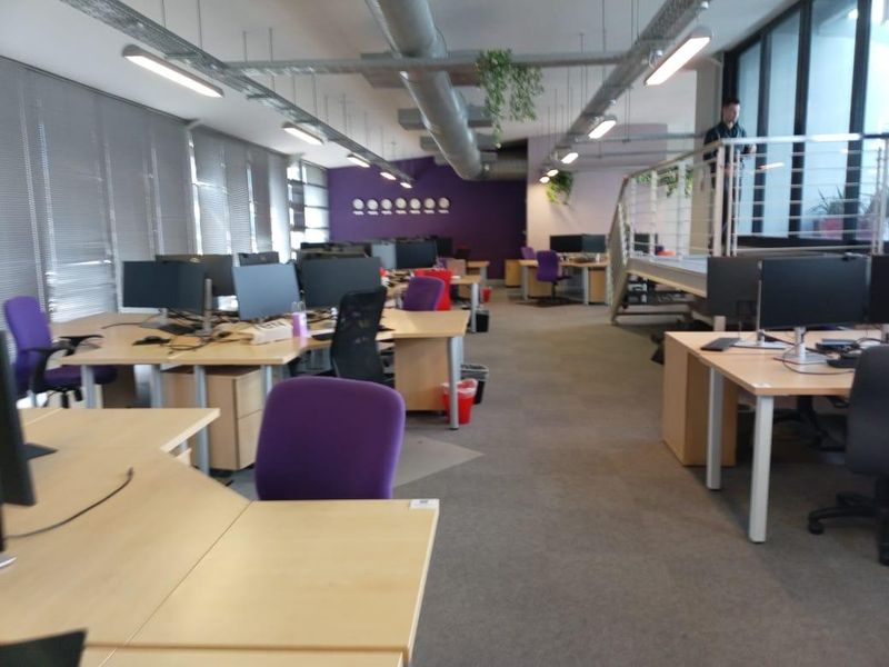 Open-plan office space featuring exposed industrial ductwork, linear lighting, and a mezzanine level with a metal staircase.
