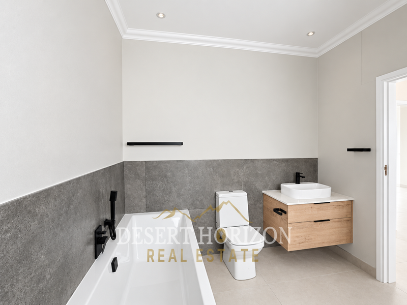 Modern bathroom featuring a built-in bathtub, wall-mounted wooden vanity with countertop basin, toilet, and contemporary black matte fixtures with neutral tiled finishes.
