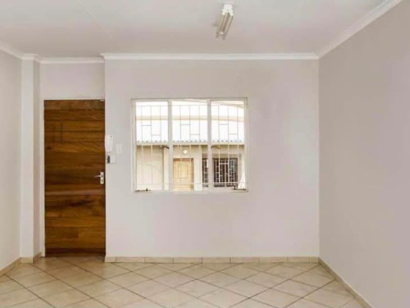 A bright, empty living area featuring neutral tiled flooring, a wooden door, and a window fitted with security bars.