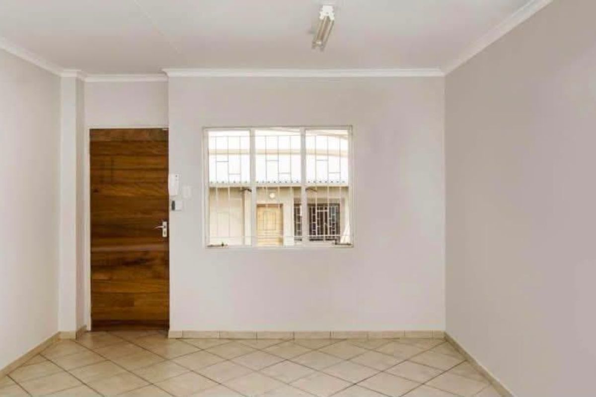 A bright, empty living area featuring neutral tiled flooring, a wooden door, and a window fitted with security bars.