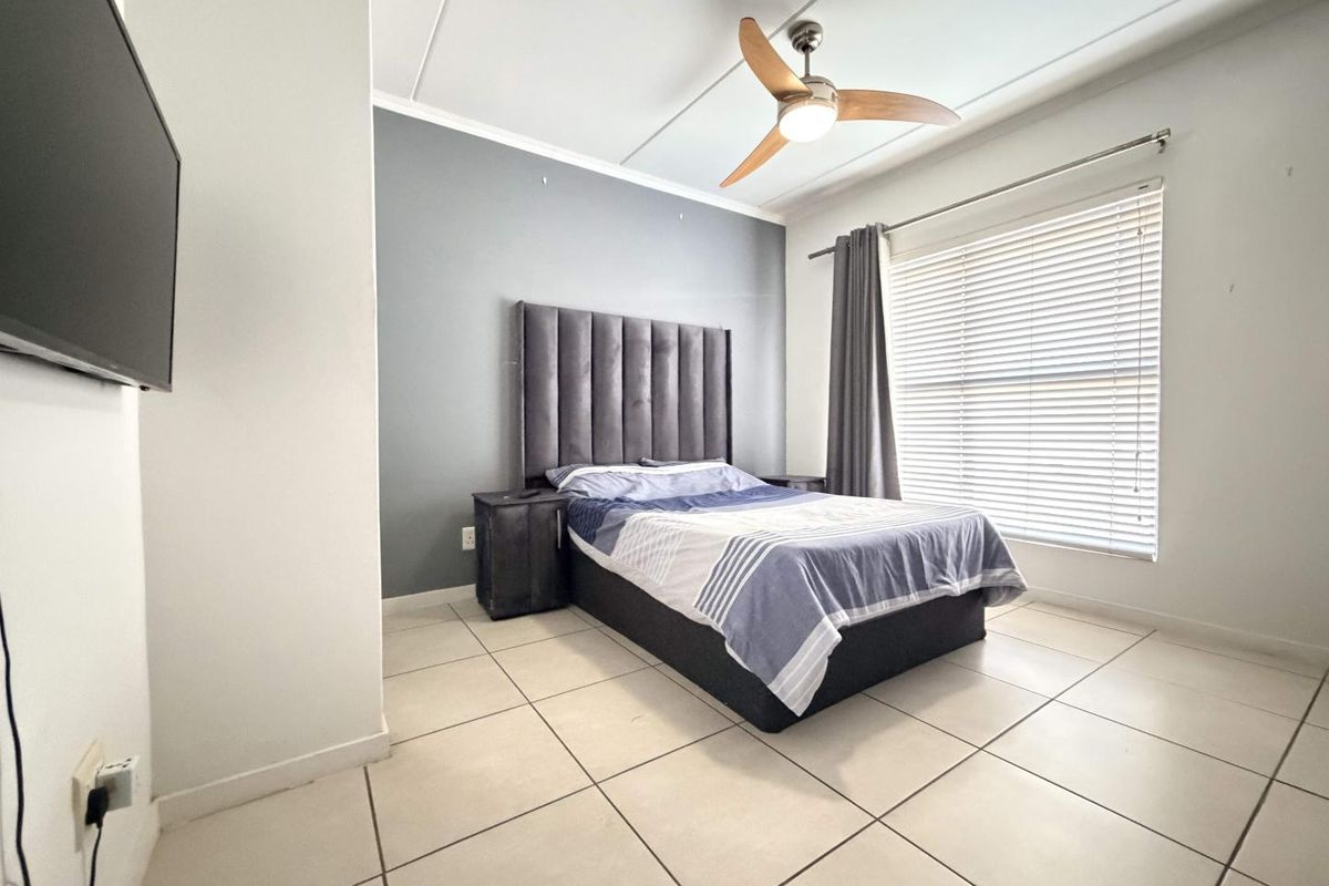 A well-lit bedroom featuring neutral tiled flooring, a modern ceiling fan, and venetian blinds, providing a clean and functional living space.