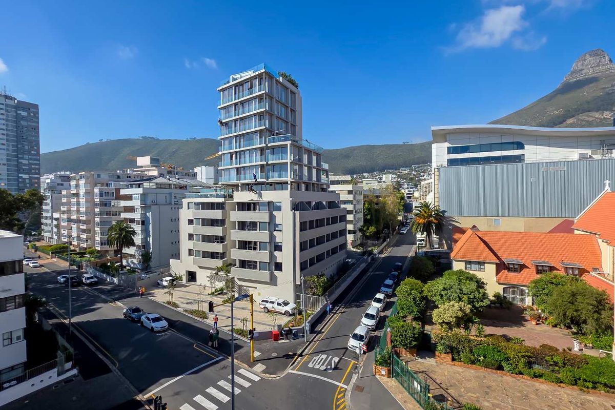 An elevated aerial view of a modern multi-story apartment building situated in a developed urban neighborhood with mountain views in the background.