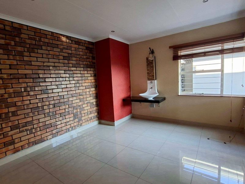 A bright bedroom featuring a prominent exposed brick wall, tiled flooring, and a modern wall-mounted basin, offering a clean and versatile living space.