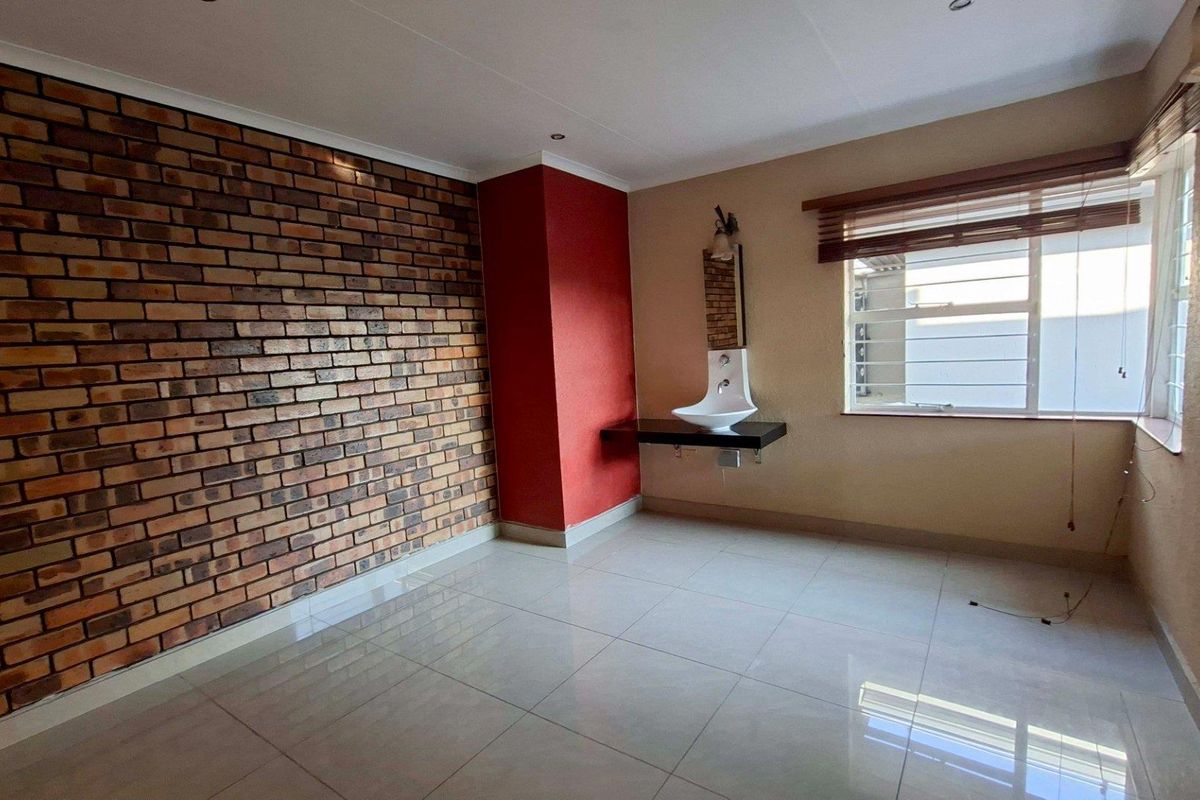 A bright bedroom featuring a prominent exposed brick wall, tiled flooring, and a modern wall-mounted basin, offering a clean and versatile living space.