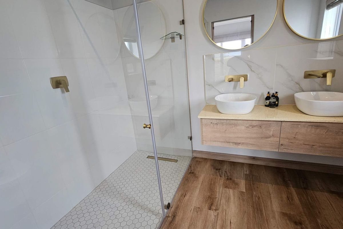 Modern bathroom featuring a floating wood-finish vanity with dual vessel sinks, gold-tone fixtures, and a glass-enclosed walk-in shower with white tiled walls.