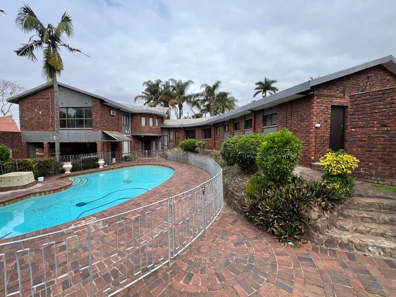 Spacious outdoor pool area featuring a curved swimming pool, brick paving, and established garden beds set against a face brick house exterior.