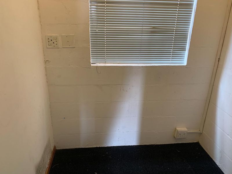 A small office space featuring exposed concrete block walls, carpet flooring, and fitted venetian blinds, showing signs of damp at the base.