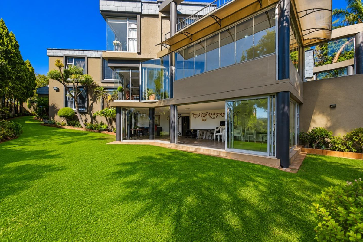 Rear view of a modern multi-story residence featuring extensive glass sliding doors, a spacious balcony, and a well-maintained landscaped garden.