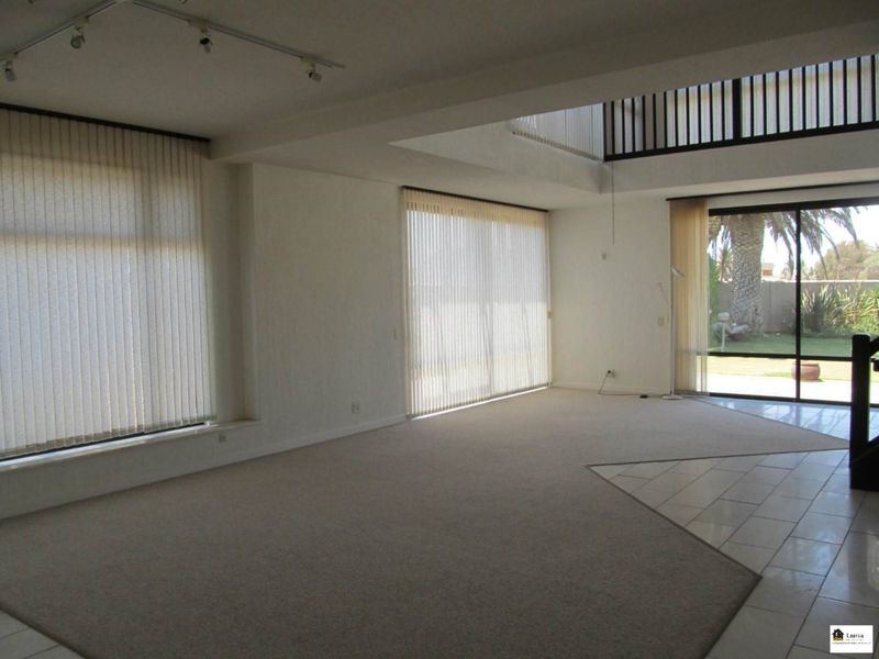 Spacious, light-filled living area featuring double-volume ceilings, a mezzanine level, large sliding glass doors, and a combination of carpeted and tiled flooring.