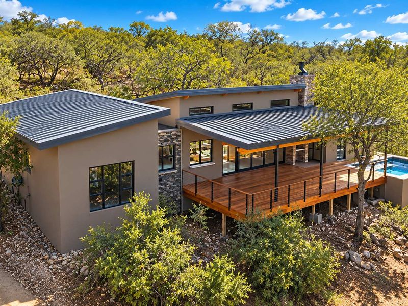 Aerial view of a modern home featuring a metal roof, expansive wooden deck, stone accents, and a private swimming pool set in a natural landscape.