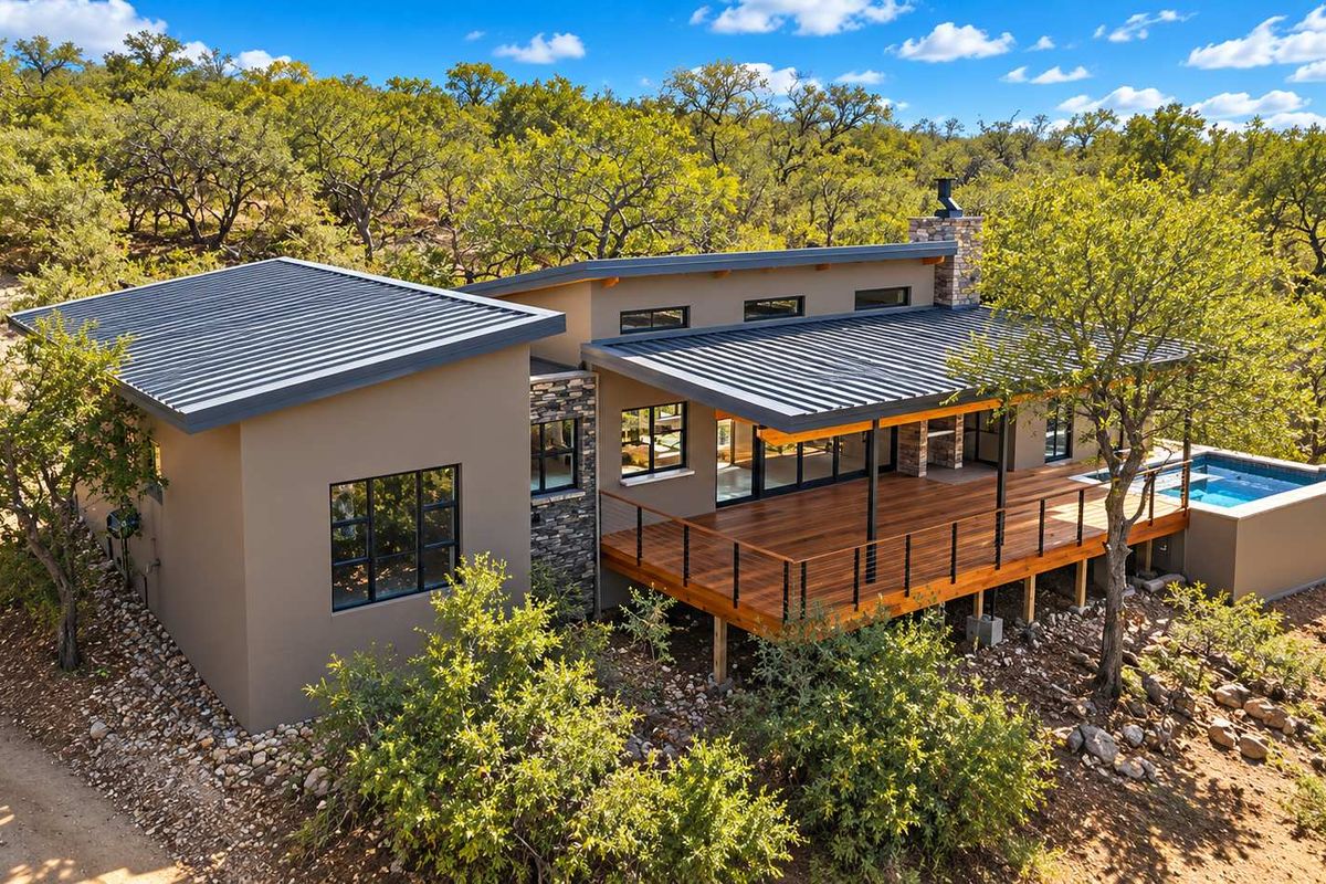 Aerial view of a modern home featuring a metal roof, expansive wooden deck, stone accents, and a private swimming pool set in a natural landscape.