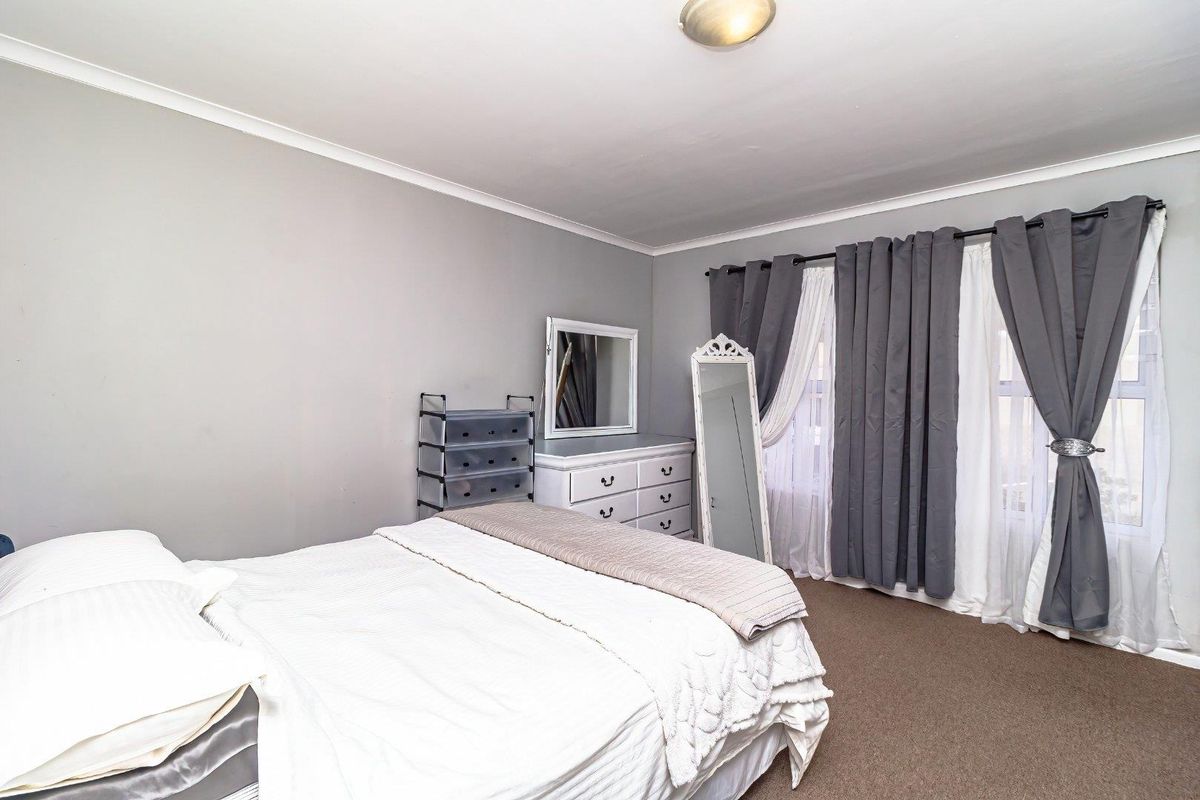 A well-lit bedroom featuring neutral wall tones, carpeted flooring, and large windows dressed with layered curtains.