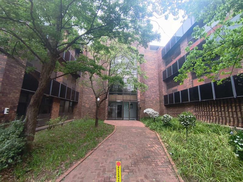 A view of the office building entrance featuring a brick facade, paved walkway, and established garden landscaping.