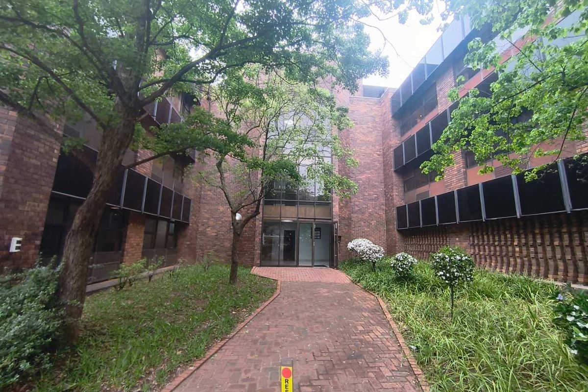 A view of the office building entrance featuring a brick facade, paved walkway, and established garden landscaping.