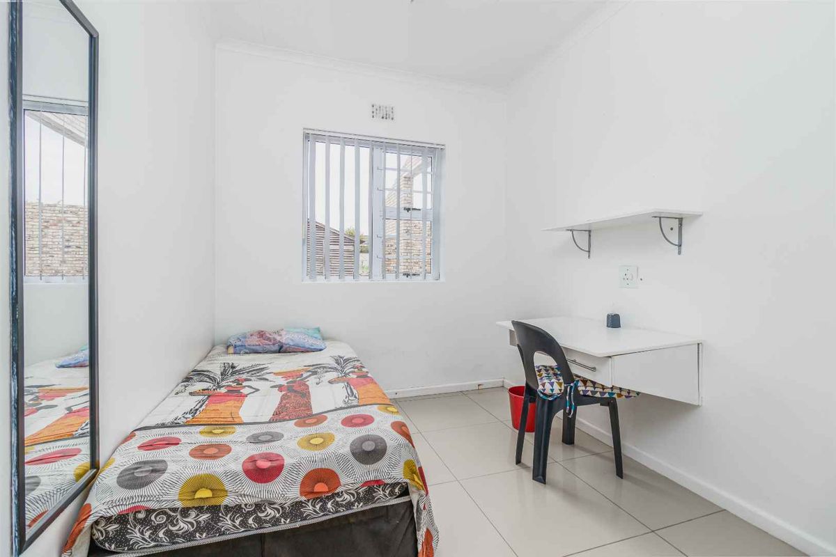 A functional bedroom featuring tiled flooring, a built-in wall-mounted desk with shelving, and a window with security bars.