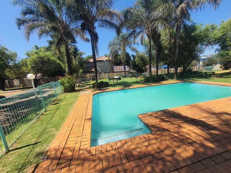 A communal swimming pool area featuring brick paving, established palm trees, and green lawn within a fenced residential complex setting.