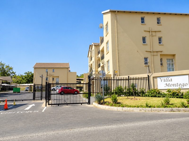 Front entrance view of Villa Montego townhouse complex featuring secure gated access, paved driveway, and multi-story residential building facade.