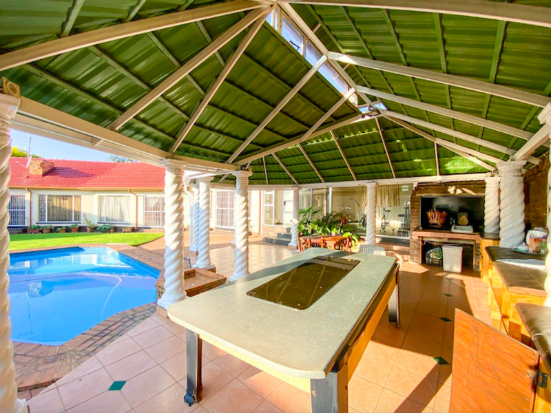 Spacious covered patio featuring a built-in braai area, tiled flooring, and decorative columns, overlooking a private swimming pool.