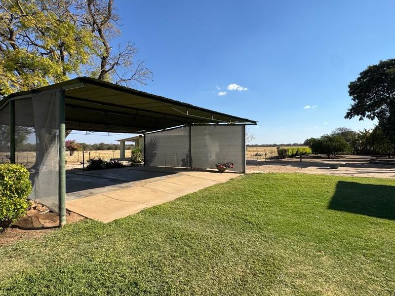 A spacious outdoor covered patio area with a metal roof structure and concrete flooring, set within a large landscaped farm garden.