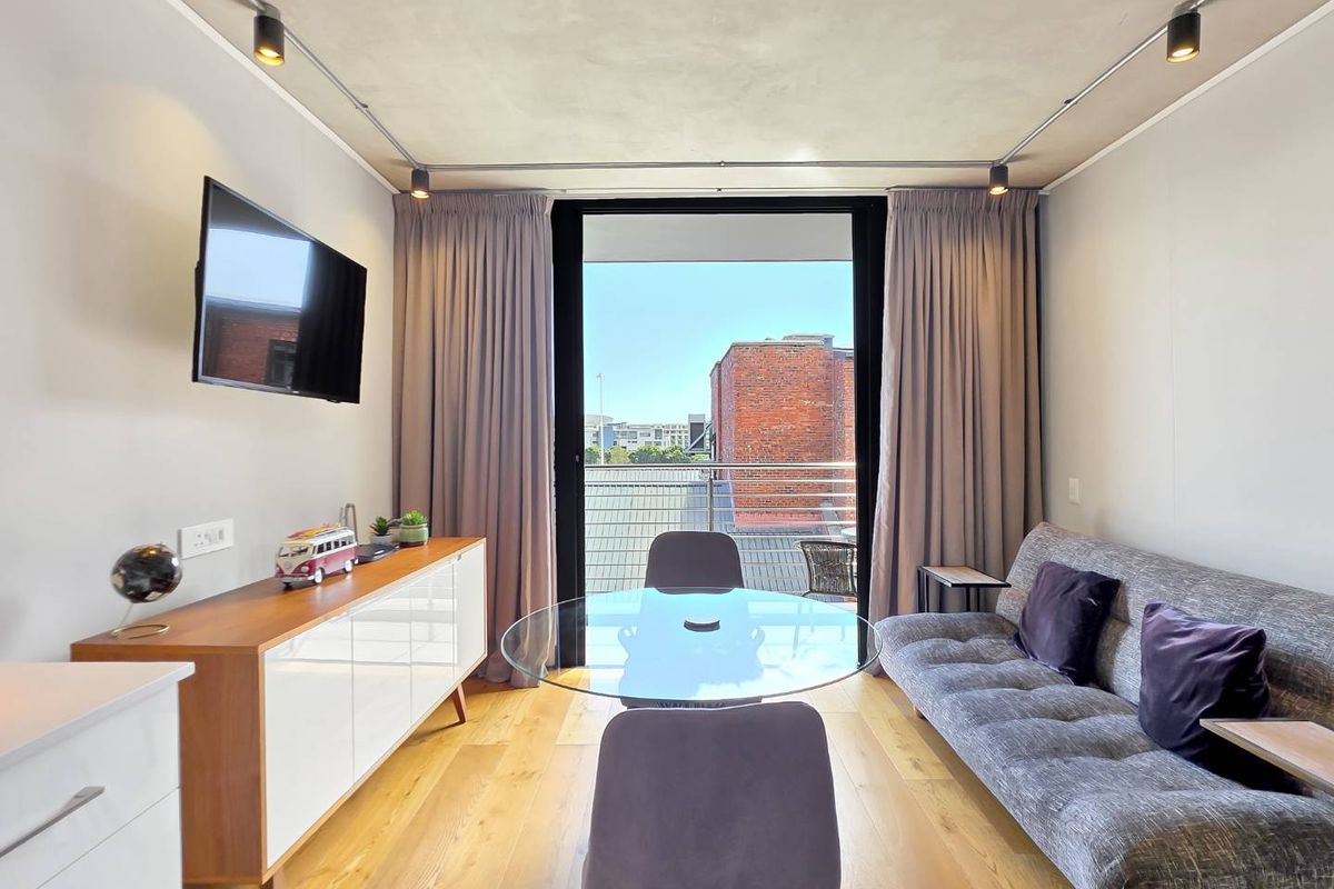 A modern living room featuring hardwood flooring, track lighting, and a large sliding glass door providing access to a balcony with urban views.