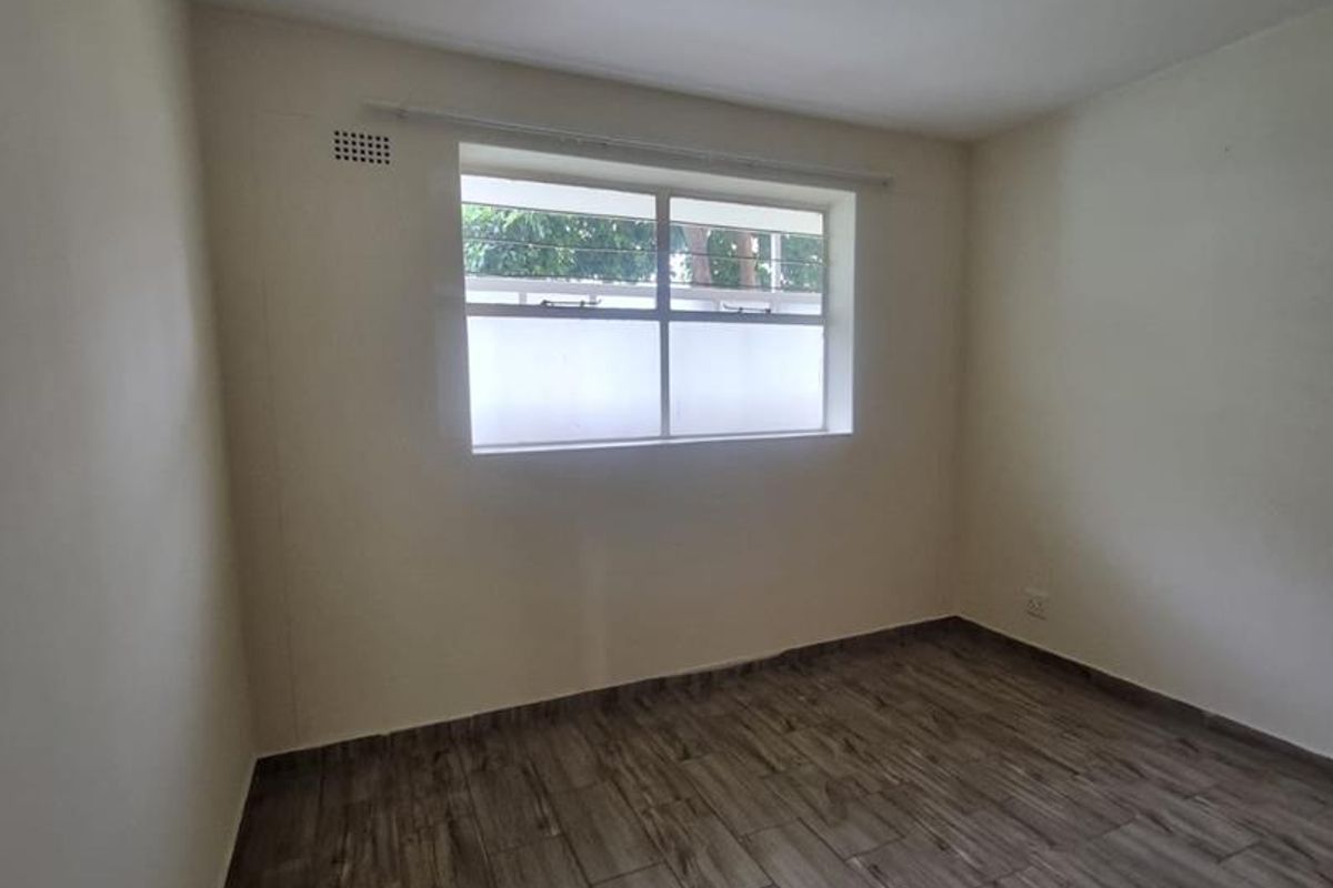 A clean, empty bedroom featuring modern wood-look tile flooring, neutral walls, and a window with security bars.