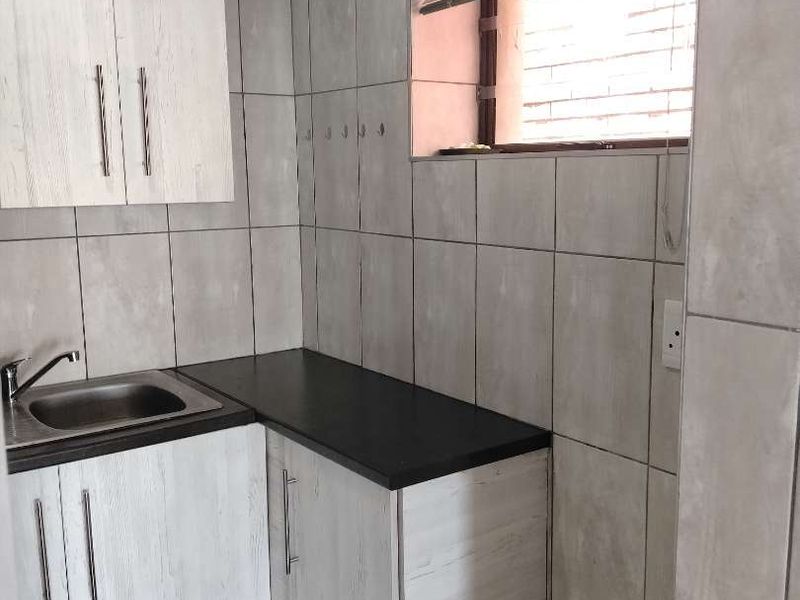 A compact kitchenette featuring light wood-grain cabinetry, a stainless steel sink, dark countertops, and full-height wall tiling.