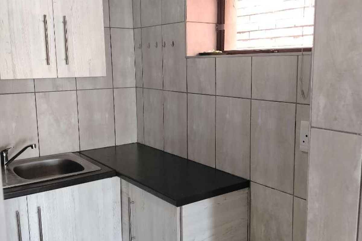 A compact kitchenette featuring light wood-grain cabinetry, a stainless steel sink, dark countertops, and full-height wall tiling.