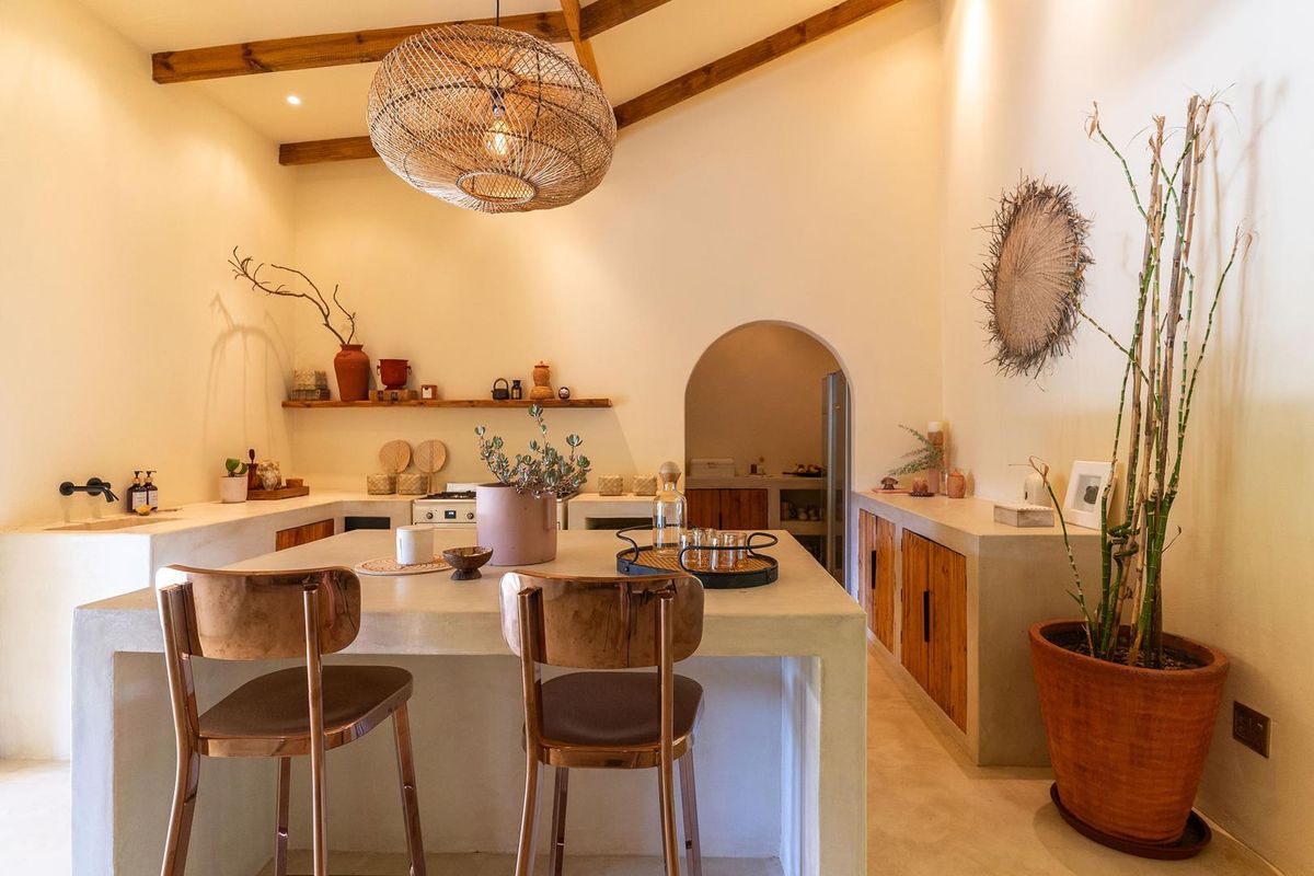 A stylish kitchen featuring a central island with concrete-style countertop, wooden cabinetry, exposed ceiling beams, and an arched doorway leading to a scullery area.