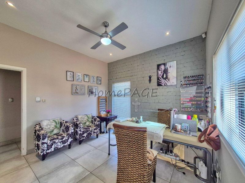 A functional office space featuring tiled flooring, a ceiling fan, and an exposed brick feature wall with natural light from venetian blinds.