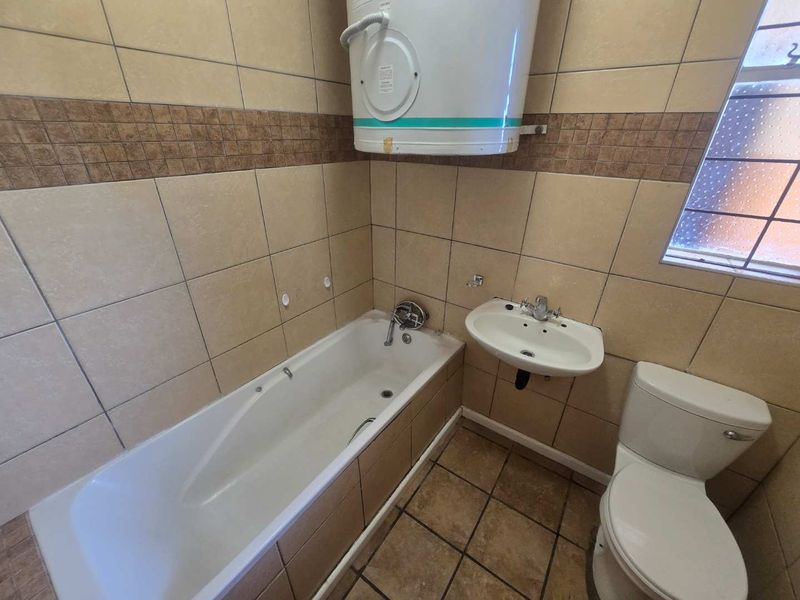 A functional bathroom featuring a bathtub, wall-mounted basin, toilet, and an electric water heater, finished with neutral wall and floor tiling.