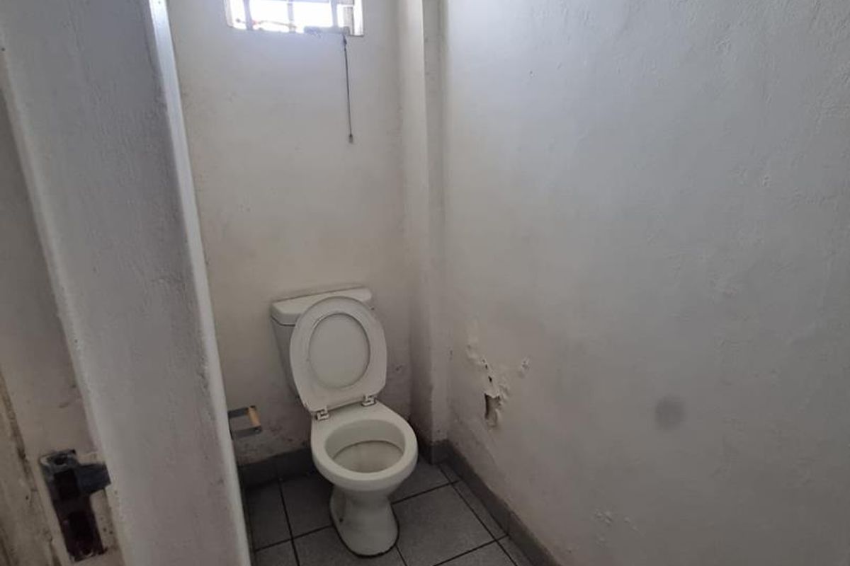 A small, functional bathroom featuring tiled flooring, a toilet, and a window with security bars, currently requiring cosmetic wall repairs.