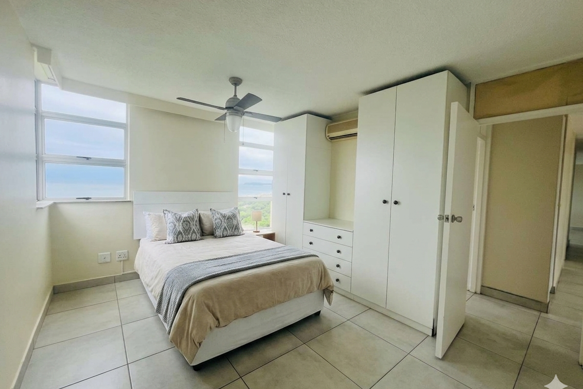 A bright bedroom featuring built-in white wardrobes, a ceiling fan, and large windows offering scenic coastal views.