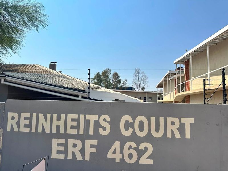 Exterior view of the Reinheits Court apartment complex entrance, featuring a boundary wall with electric fencing and visible building roof structures.