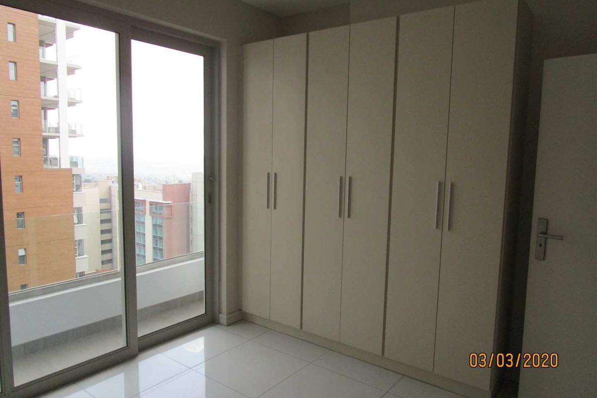 A bright bedroom featuring large sliding glass doors leading to a balcony and floor-to-ceiling built-in wardrobes with a neutral finish.