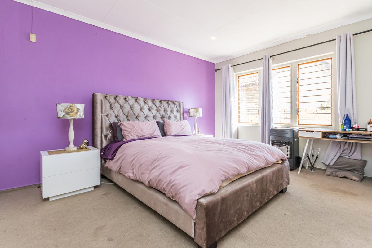 A spacious bedroom featuring a prominent purple accent wall, large windows with fitted blinds, and neutral carpeting.